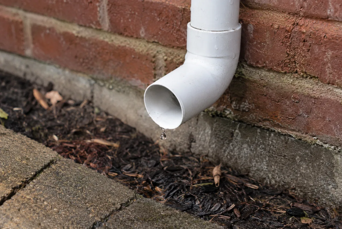 White PVC condensate drain pipe running down a brick wall, discharging a small drip of water into mulch below during normal air conditioner operation