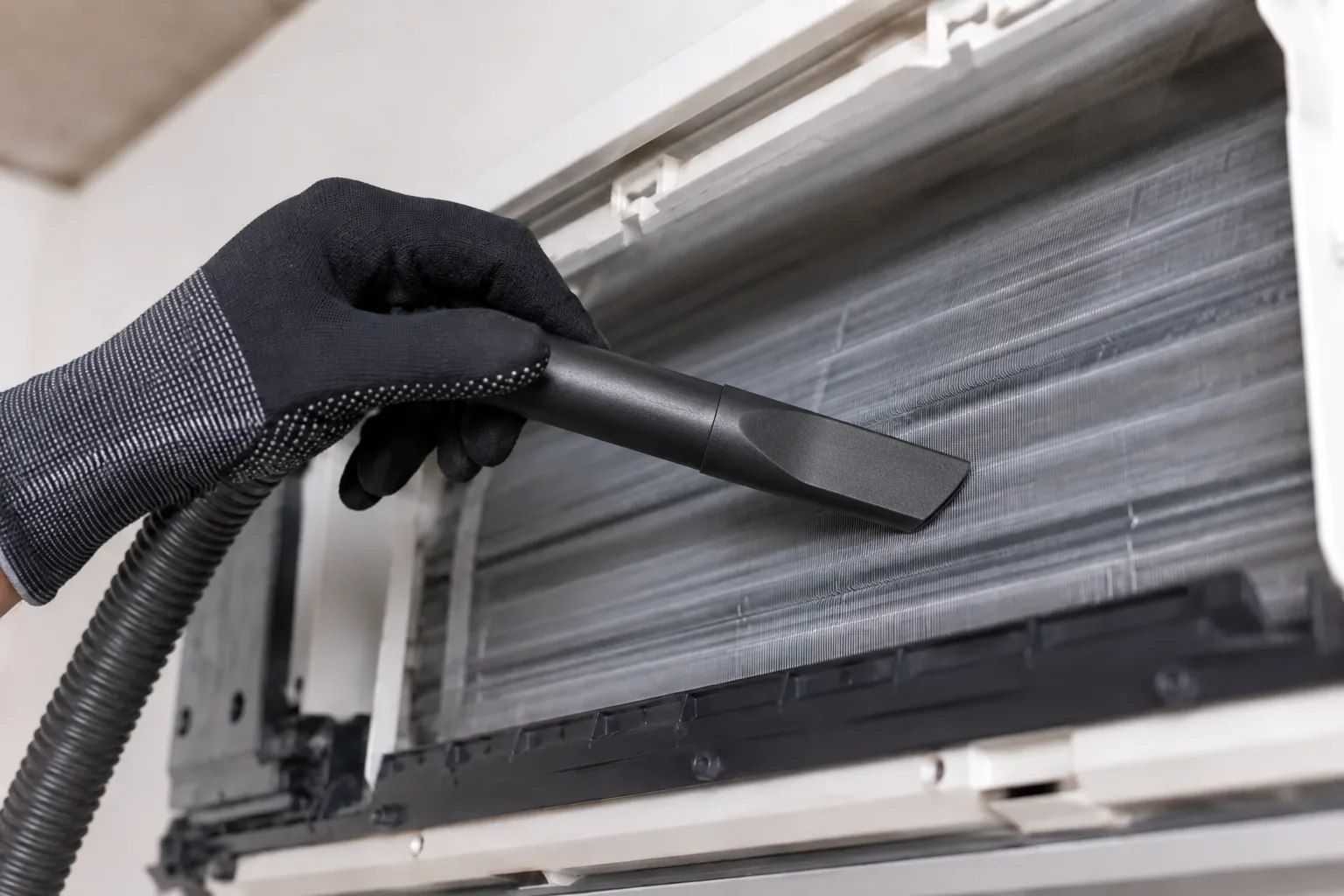 Gloved hand using a slim vacuum attachment to clean dust from the evaporator coil fins of a wall-mounted split system with the front panel and filters removed