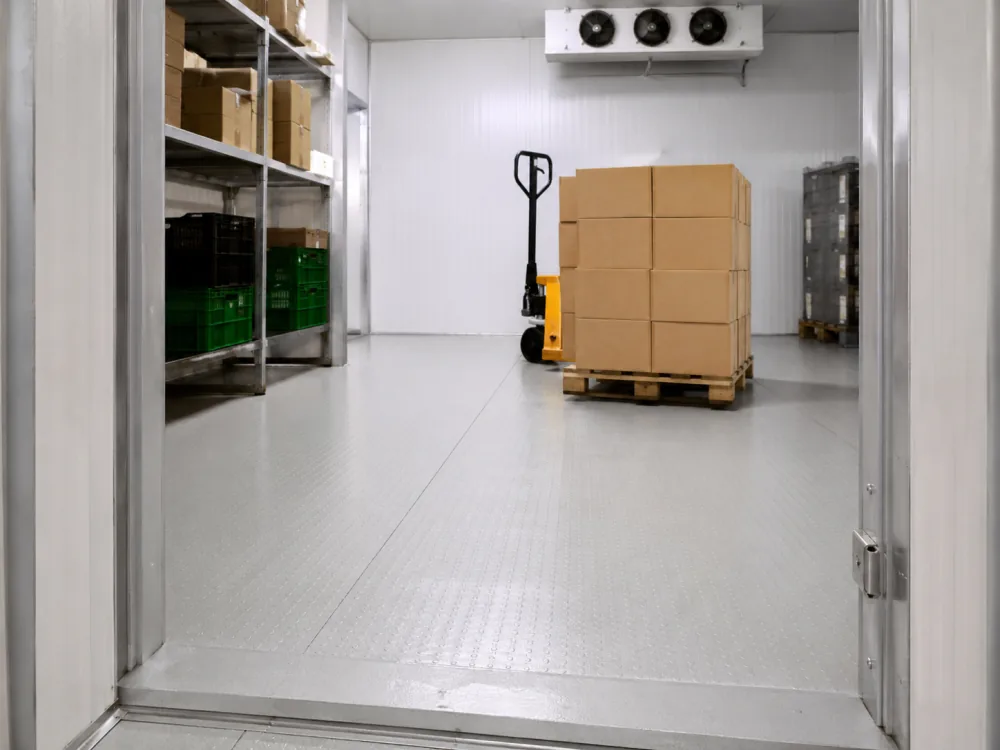 Inside a commercial cool room with a pallet jack, cardboard boxes on a pallet and shelved crated produce, showing how floor traffic and loading equipment shape the design