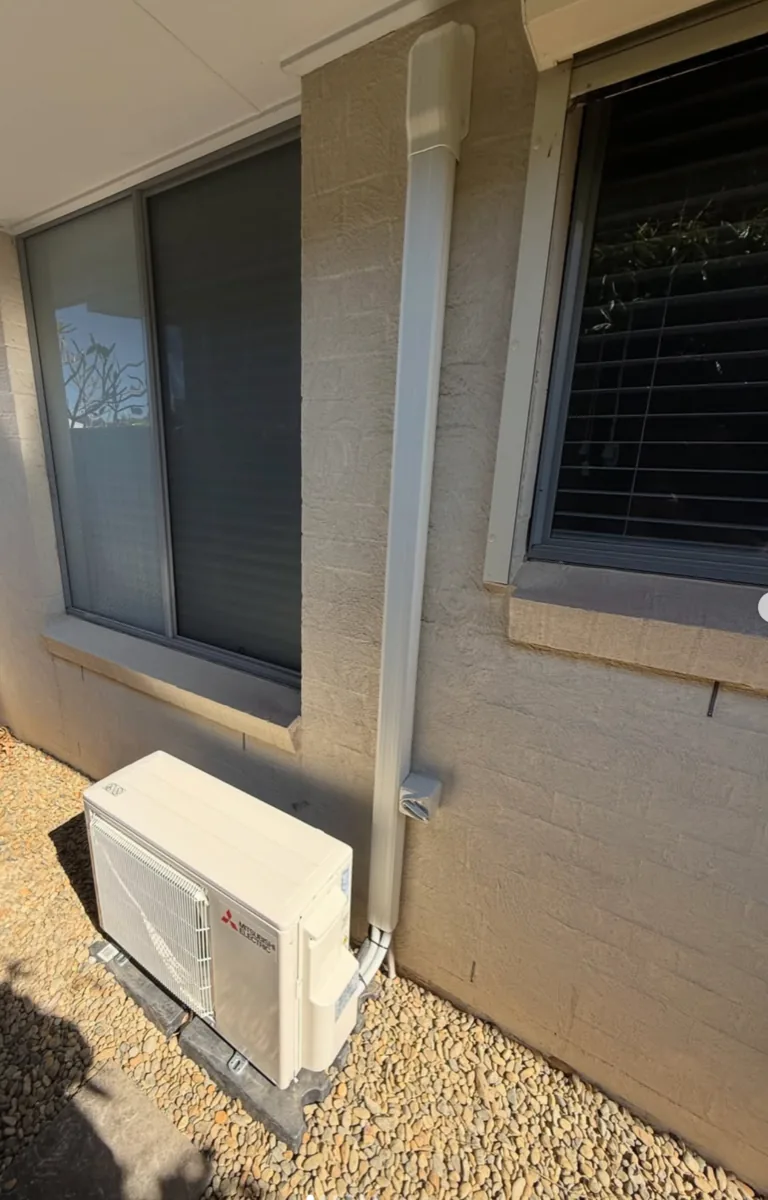Mitsubishi Electric split system outdoor condenser installed on a paved area against a beige rendered wall, with the refrigerant line concealed inside a white line-set cover running up to the eave